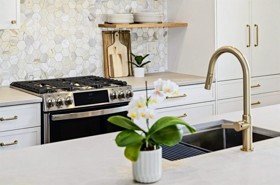 A modern kitchen featuring a stainless steel gas range with a hexagonal tile backsplash in neutral tones. White cabinetry with gold hardware surrounds the area, and an open wooden shelf holds neatly stacked plates.